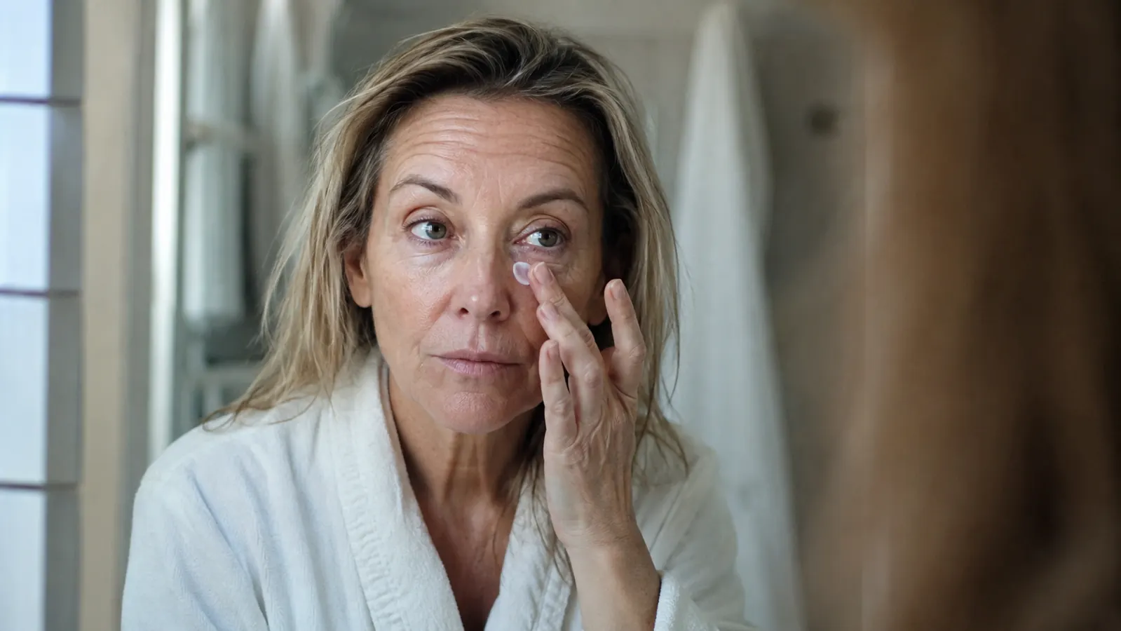 A woman in her fifties applying eye cream with her ring finger under her eye at a bathroom mirror in the morning
