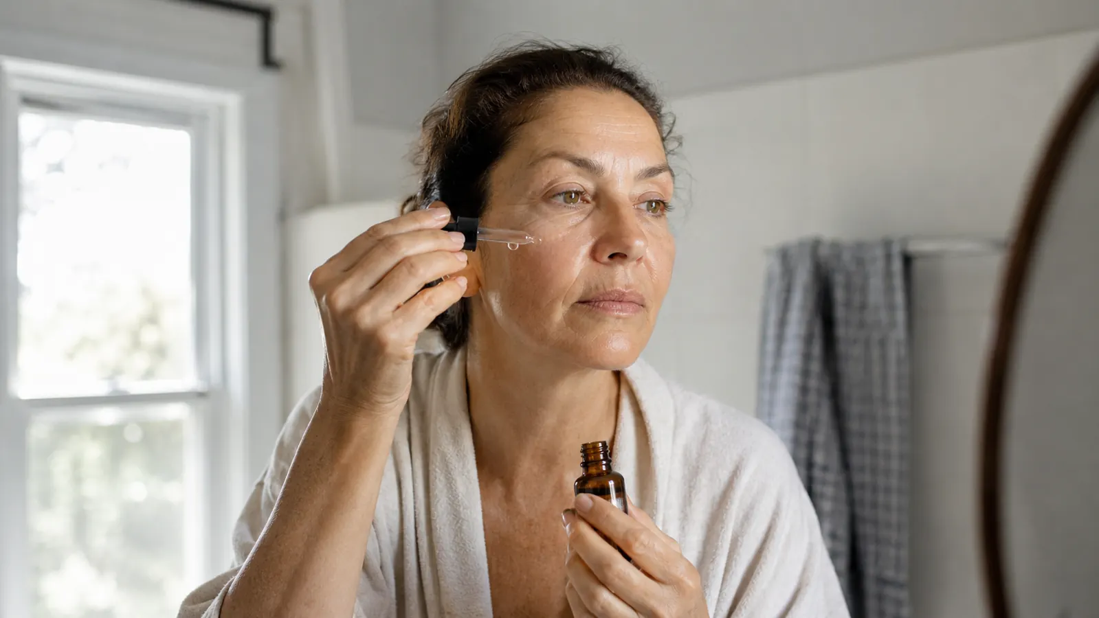 A woman in her fifties applying a few drops of vitamin C serum to her face at a bathroom sink in soft morning light