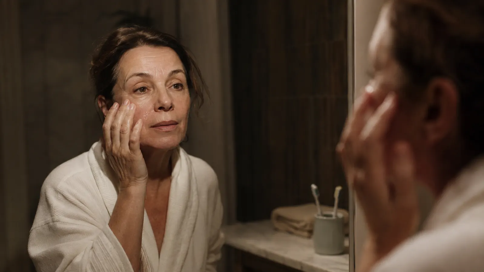A woman in her fifties stands at a bathroom sink at night, pressing a small amount of lotion into her cheek under warm, dim lighting