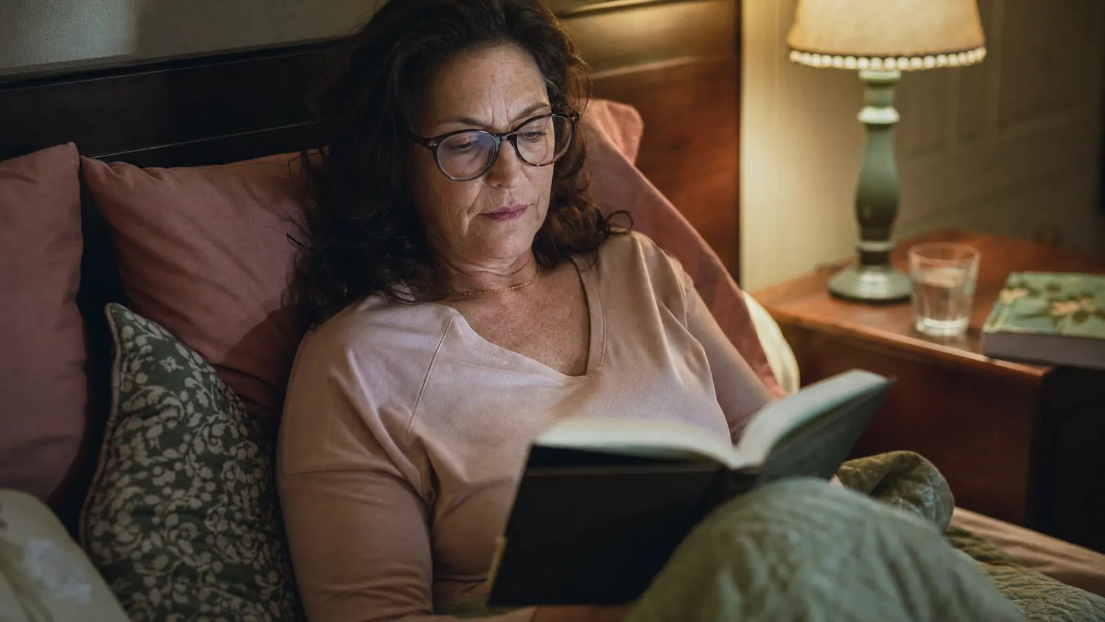 A woman in her fifties reading in bed by a small lamp, nightstand visible with a folded book and a glass of water
