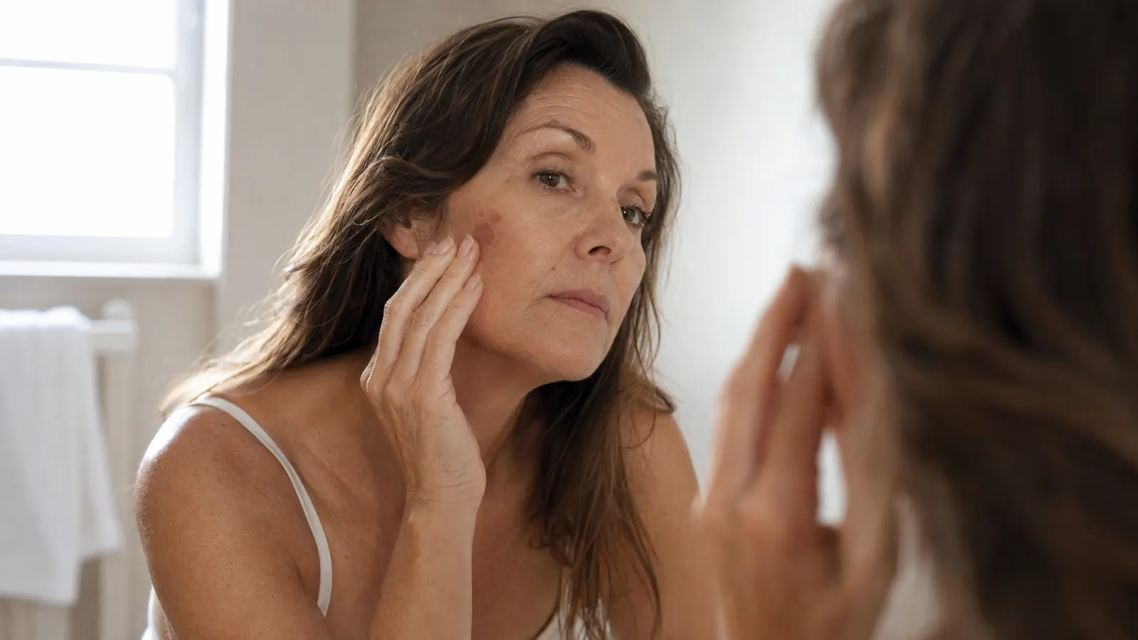 A woman in her fifties standing close to a bathroom mirror in early morning light, studying a dark spot near her cheekbone with a quiet, thoughtful expression