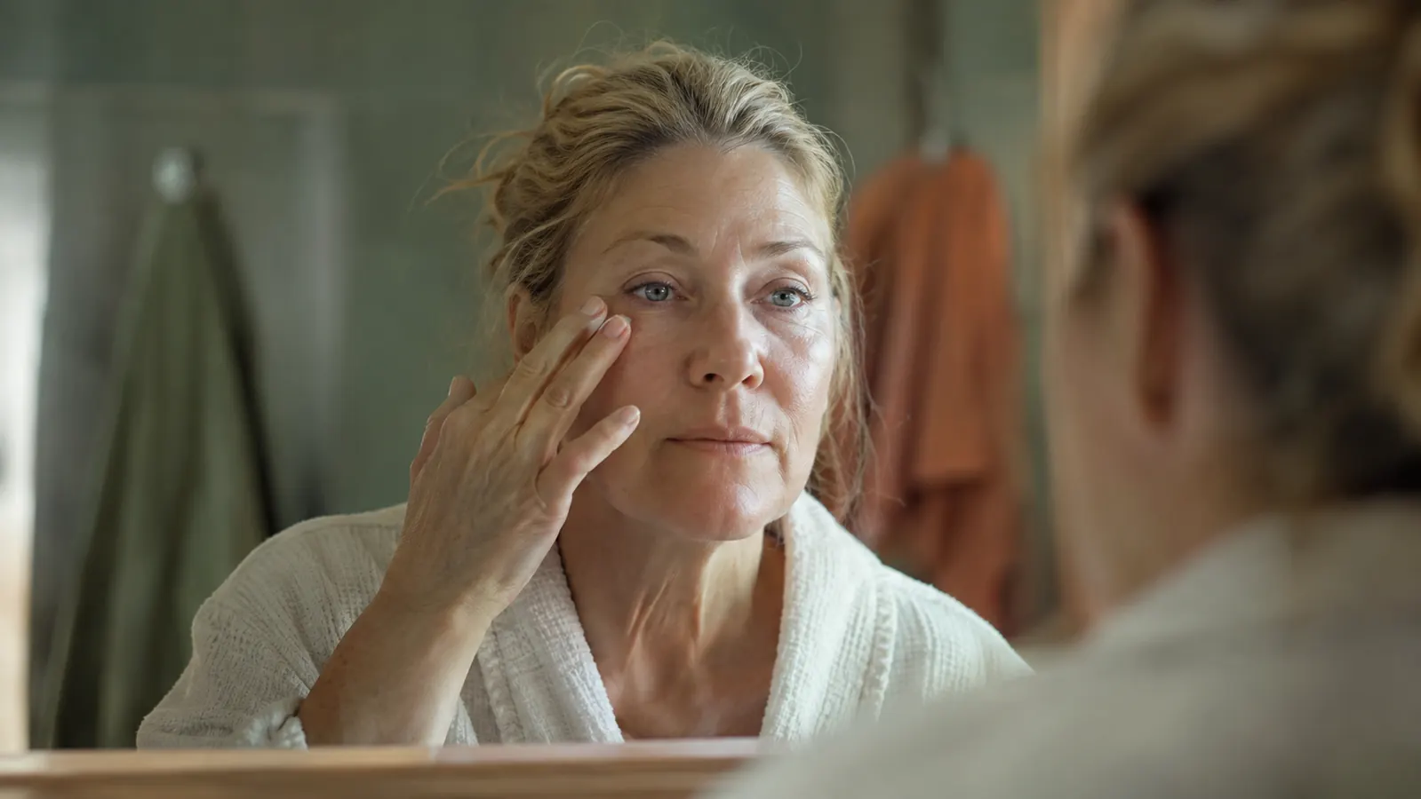 A woman in her fifties at a bathroom mirror in soft morning light, gently pressing a fingertip under one eye